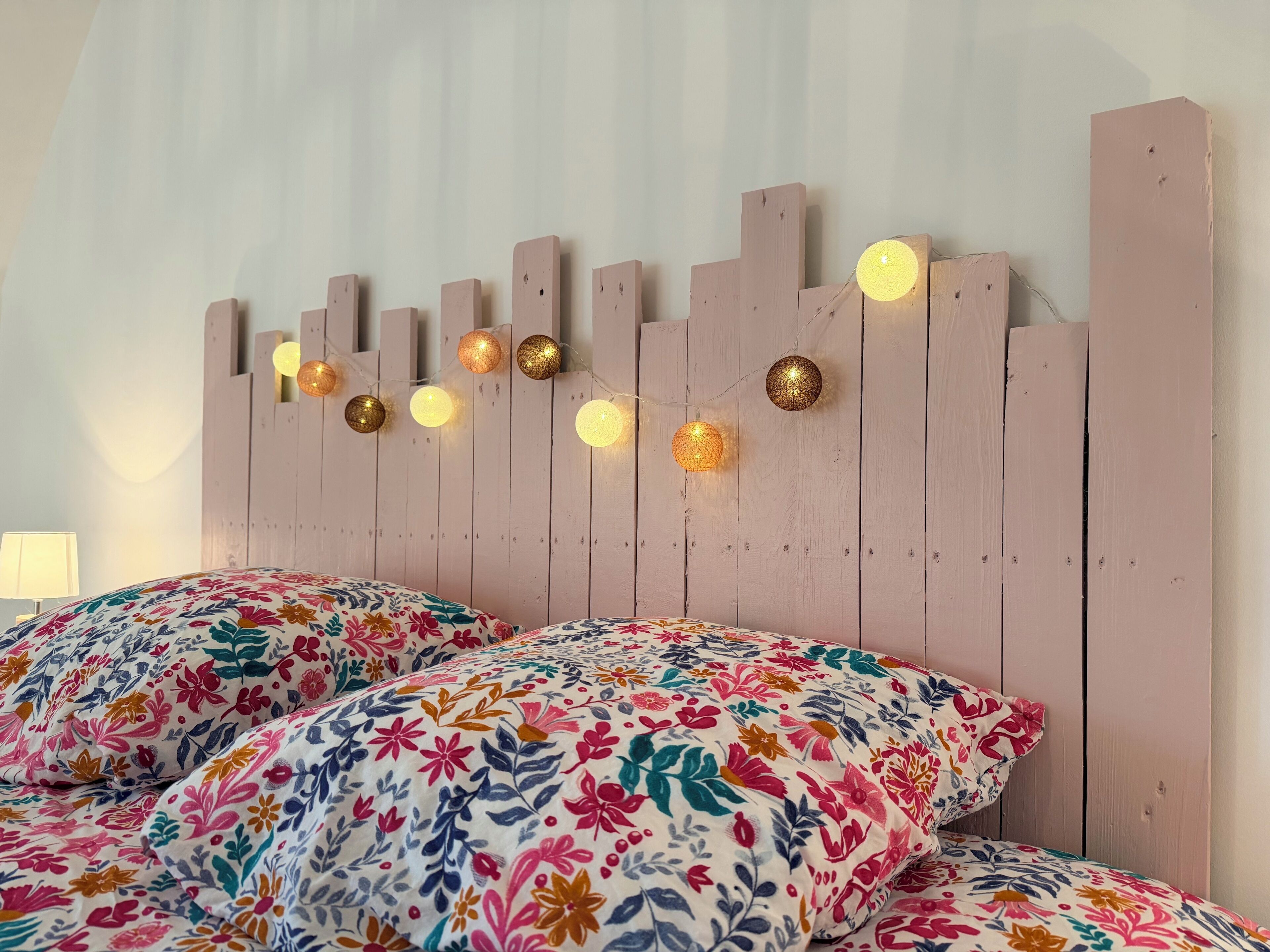 Un lit avec des oreillers à motifs floraux et une tête de lit en bois décorée de guirlandes lumineuses.
