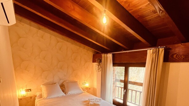 Een slaapkamer met een houten plafond, een bed met witte lakens, een bankje en een raam met gordijnen.