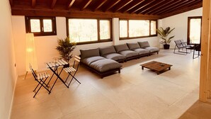 Een ruime woonkamer met een hoog houten plafond, een hoekbank, een salontafel en een eethoek met stoelen.