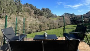 Un patio con dos sillones negros y una mesa, con vista a un paisaje verde con colinas y un cielo azul despejado.