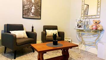 Un salon avec deux fauteuils sombres, une table basse en bois, une petite table d’appoint sur laquelle est posé un ours en peluche, un tableau encadré au mur et un miroir décoratif.