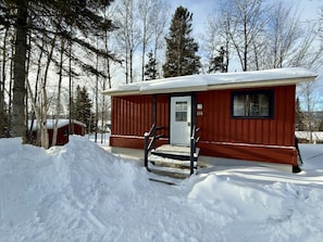 A cabin with a red exterior, a white door, and a small porch with steps leading up to it.