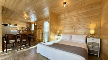 Interior de una cabaña de madera con cama, comedor y pequeña cocina.