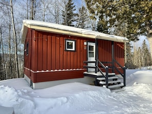 Una cabaña roja con puerta blanca y un ventanal pequeño, rodeada de nieve y árboles.