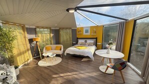 A modern hotel room with a large bed, a sofa, a small table, and a chair. There is a large window providing a view of the outside. - Unusual gîte with spa & sauna - 15 min from Mont-St-Michel ✨ the starry sky✨ (Juilley)