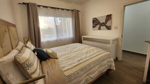 Une chambre à coucher comprenant un lit, une table de chevet, un ventilateur de plafond, une fenêtre avec des rideaux et une porte.