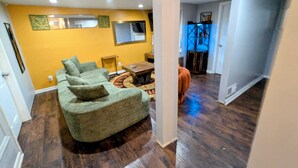 Una sala de estar con un sofá modular, una mesa de centro y una alfombra. Hay una ventana con persianas, un espejo en la pared y una puerta que da a otra habitación.