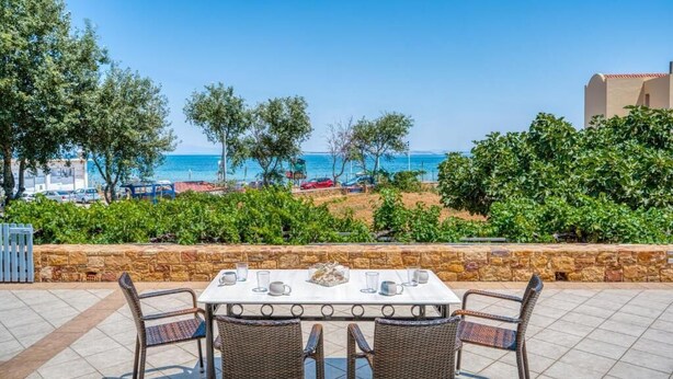 Un espace de restauration en plein air avec une table dressée pour quatre personnes, donnant sur une plage parsemée de parasols et bordée par une route où circulent des voitures.