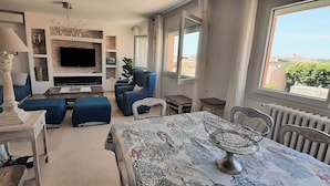 A modern living room with a flat-screen TV, blue upholstered seating, a dining table with a patterned tablecloth, and a view of the city through large windows. - Very Nice 3-Room Apartment, 84 m2, Spacious, Saint-Maxime, Beach 300 Meters Away (Sainte-Maxime)
