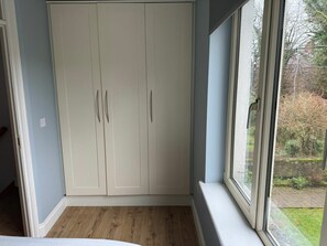 A room with a white wardrobe, a wooden floor, and a window with a view of trees and a building.