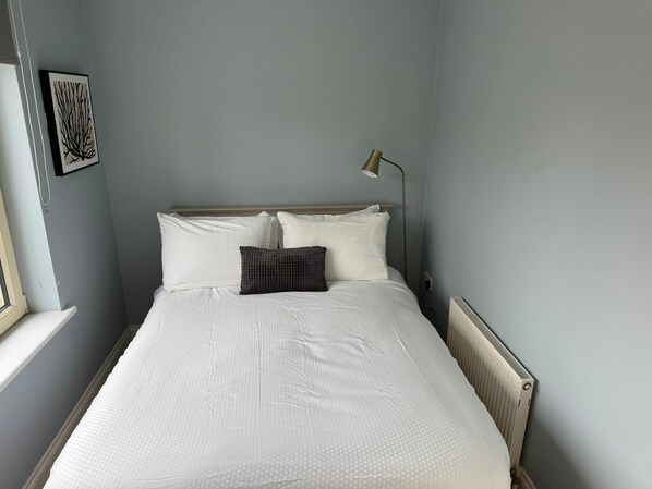 A neatly made bed with white bedding, a single dark pillow, and a bedside lamp.