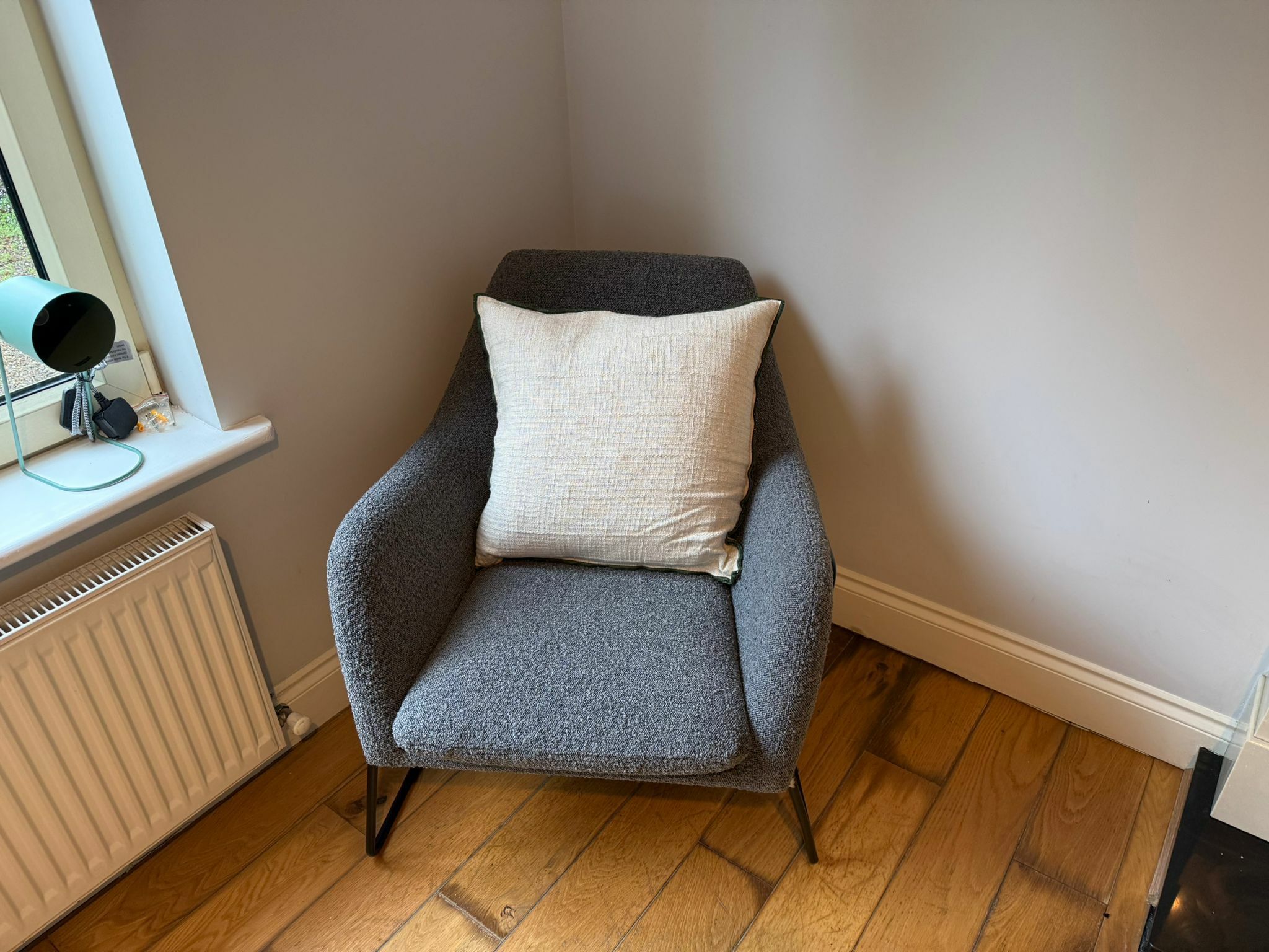 Une pièce d’angle avec un fauteuil gris, un coussin blanc, un radiateur et une fenêtre équipée d’une caméra de surveillance.