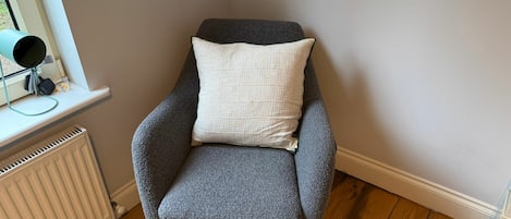 A corner room with a grey armchair, a white cushion, a radiator, and a window with a security camera.