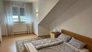 A bedroom with a wooden bed, patterned bedding, a nightstand with a lamp, a window with curtains, and a radiator. - Lovely and Peaceful, Cosy Guest House in Cologne (Köln)