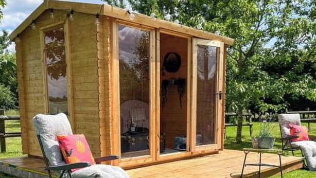 Una cabaña de madera con terraza, puerta corrediza de vidrio y dos sillones con cojines.