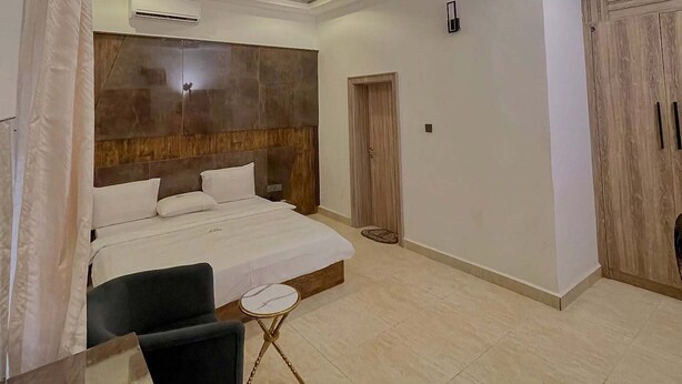 A modern hotel room with a large bed, a small desk, a chair, and a wardrobe. - Bridge Waters Hotel  (Abuja)