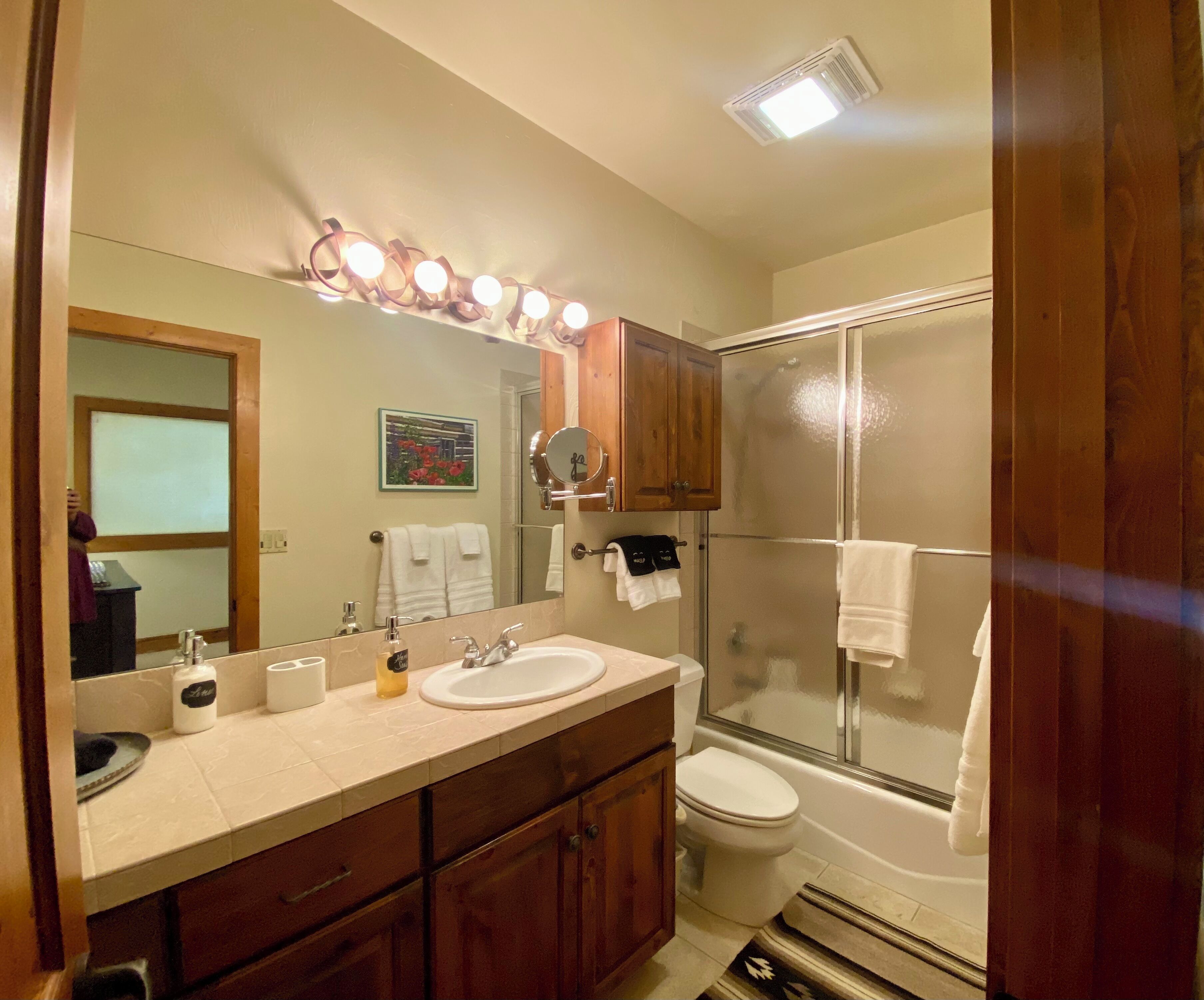 Een badkamer met een dubbele wastafel, toilet, douche met glazen deur, houten kastjes en een spiegel met verlichting.
