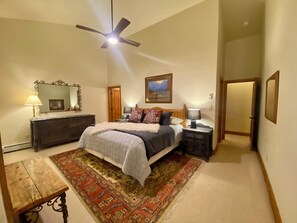 Une chambre à coucher comprenant un lit, un ventilateur de plafond, une commode, une table de chevet, une lampe, un miroir, une porte et un tapis.