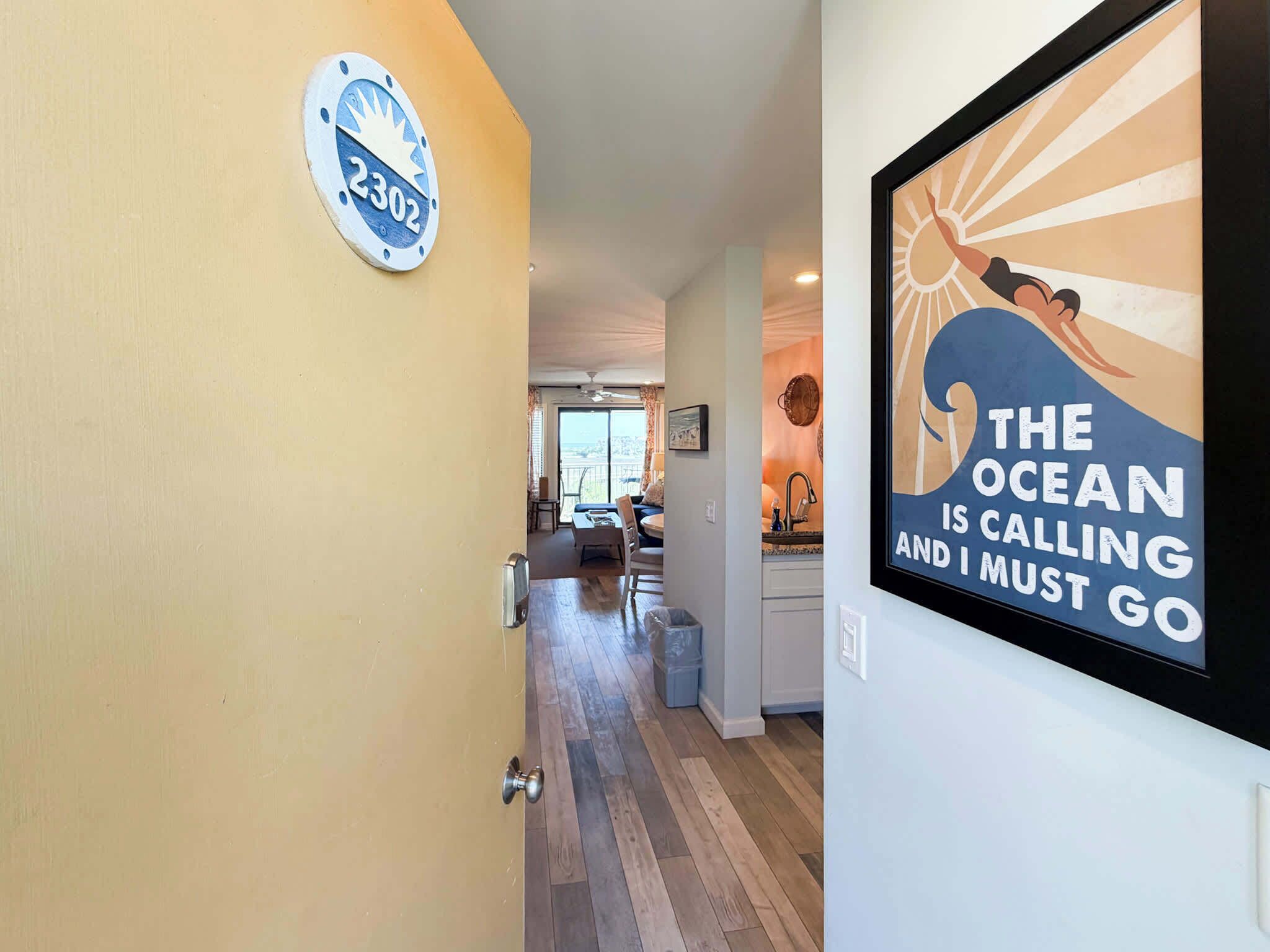 A hallway with a door marked '2302,' a framed poster of a person with a wave, and a view into a living area with a balcony.