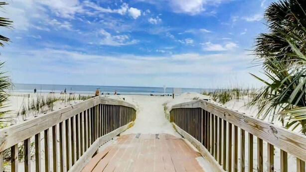 Ein hölzerner Steg führt zu einem Strand bei strahlendem Himmel.