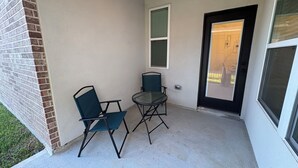 Uma pequena área de estar ao ar livre com duas cadeiras e uma mesa, adjacente a um edifício com parede de tijolos e uma porta com janela.