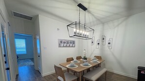 Uma sala de jantar com uma mesa de madeira posta para seis pessoas, rodeada por cadeiras. Há um lustre pendurado sobre a mesa.