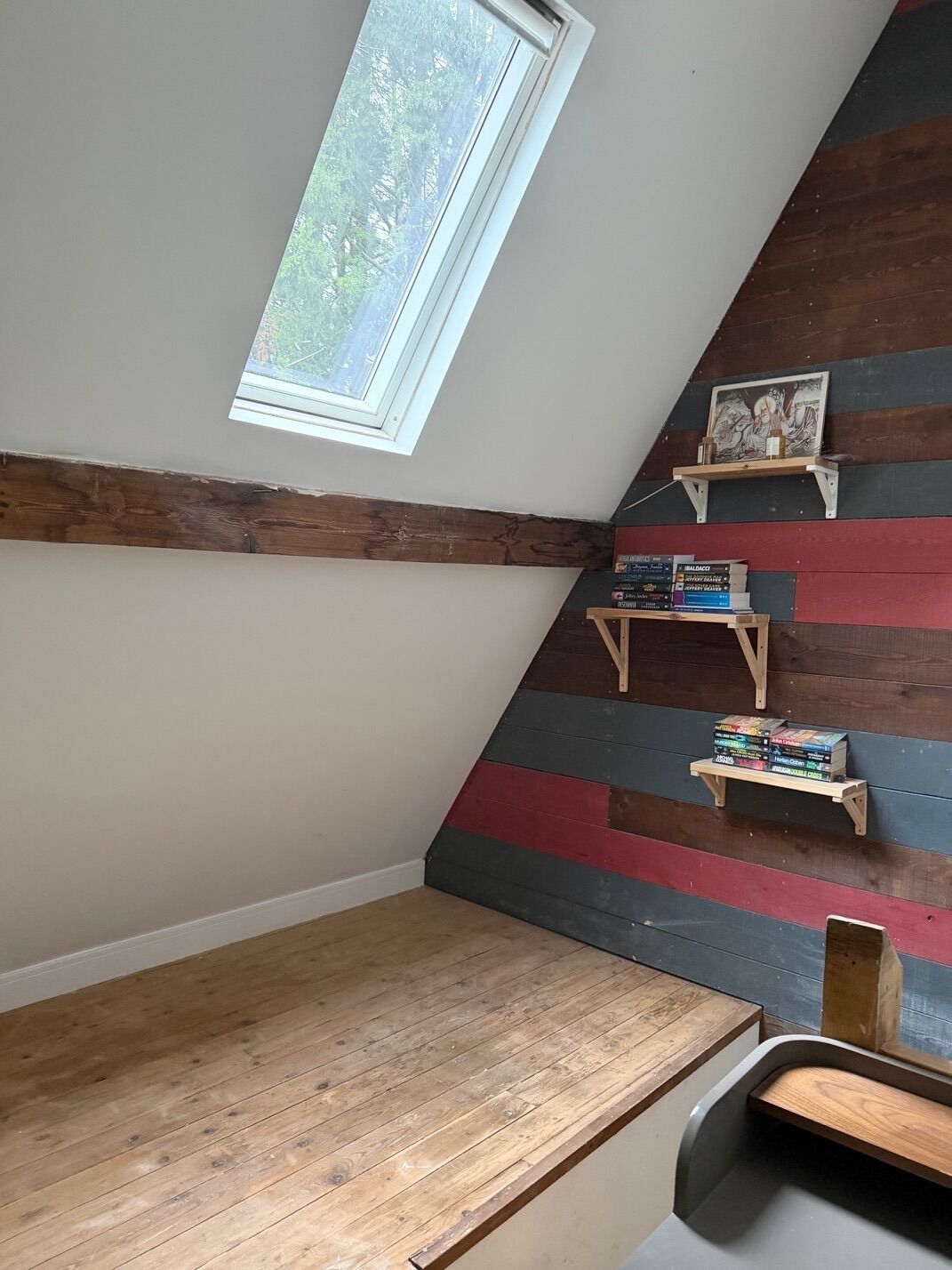 A room with a sloped ceiling, a skylight, and a wooden floor. There are two wooden shelves with books and a framed picture.