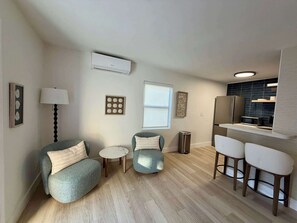 Un salon moderne comprenant un canapé, deux fauteuils, une petite table ronde et un coin cuisine avec un four à micro-ondes et des tabourets de bar.