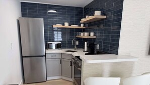 Eine kompakte Küche mit einem Edelstahl-Kühlschrank, einer Mikrowelle, einer Kaffeemaschine und Holzregalen über der Arbeitsfläche. Zwei weiße Stühle stehen an der Theke.