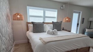 Ein ordentlich bezogenes Bett mit weißen Leinen, zwei grauen Kissen und einer beigen Tagesdecke. Darüber hängen zwei gewebte Pendelleuchten.