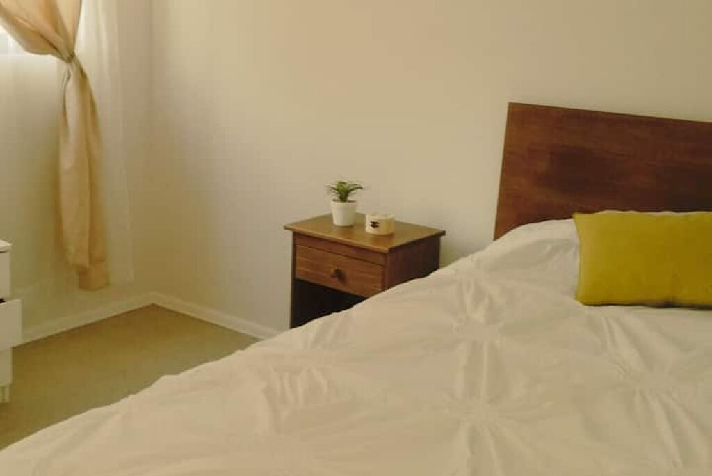 A bedroom with a bed, a nightstand, a small table with a plant, and a window with curtains.