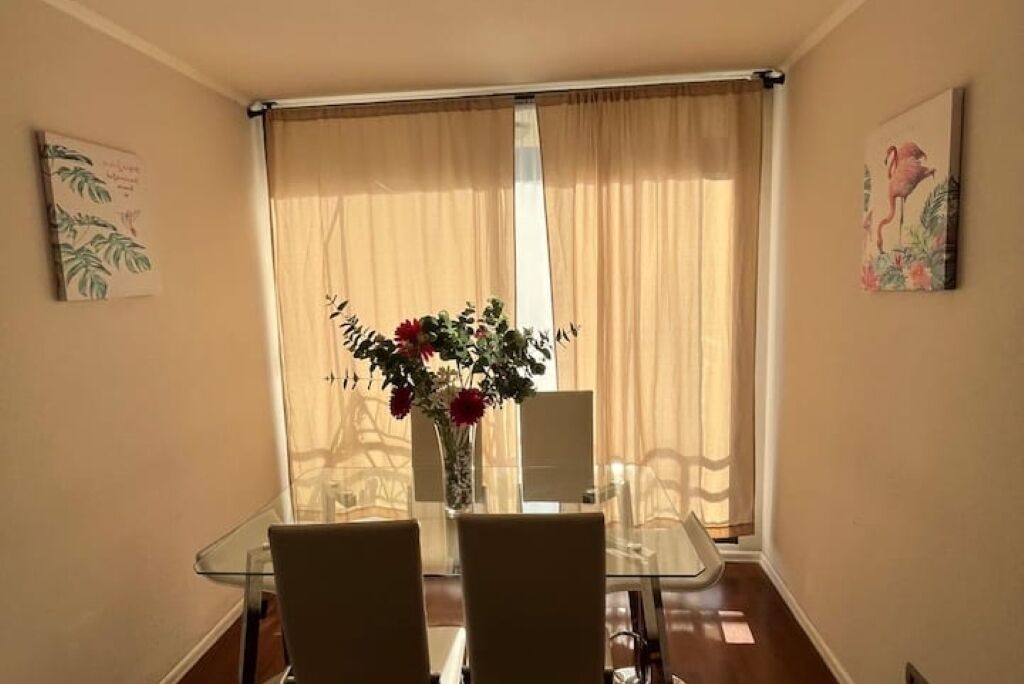 Un comedor con una mesa de vidrio y sillas, un florero con flores y vistas a través de cortinas translúcidas.