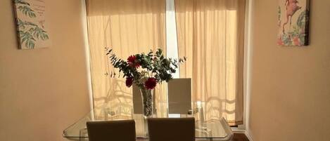 Un comedor con una mesa de vidrio y sillas, un florero con flores y vistas a través de cortinas translúcidas.