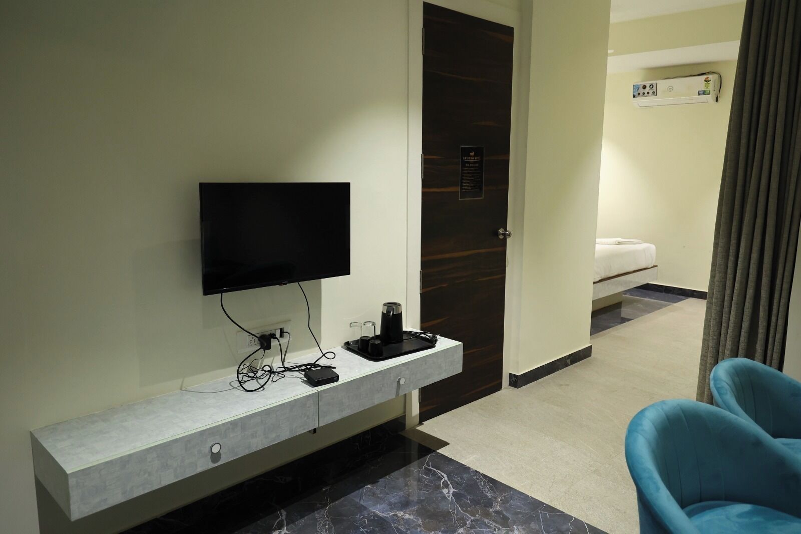 Ein Hotelzimmer mit Flachbildfernseher an einer Marmorablage, einer dunkelholzigen Tür, einem Bett mit weißer Bettwäsche und einem blauen Sessel.
