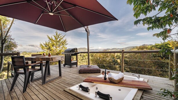 Una terraza de madera con un jacuzzi, una mesa con sillas y un paraguas grande.