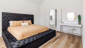 A bedroom with a black tufted headboard, a beige bedspread, a white desk with a round mirror, and a small stool. - Wonderful 4-bedroom house with AC in tranquil Houston neighborhood (Houston)