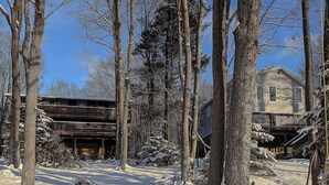A snow-covered outdoor area with a wooden deck and a stone structure, surrounded by tall trees. - Two Adjacent Lakefront Homes w/Docks, Boats, Firepit – Sleeps 24 (Pocono Lake)
