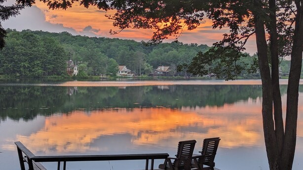 A wooden dock with two chairs and a grill, overlooking a calm lake with a forested shoreline. - Two Adjacent Lakefront Homes w/Docks, Boats, Firepit – Sleeps 24 (Pocono Lake)