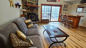 A living room with a stone fireplace, a wooden coffee table, a gray sofa with patterned cushions, and a bar area with wooden chairs. - Two Adjacent Lakefront Homes w/Docks, Boats, Firepit – Sleeps 24 (Pocono Lake)