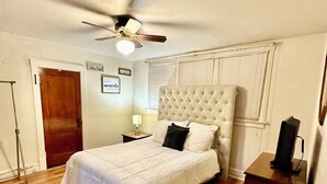 Un dormitorio con cama, ventilador de techo, piso de madera, alfombra roja, mesita de noche y cómoda.
