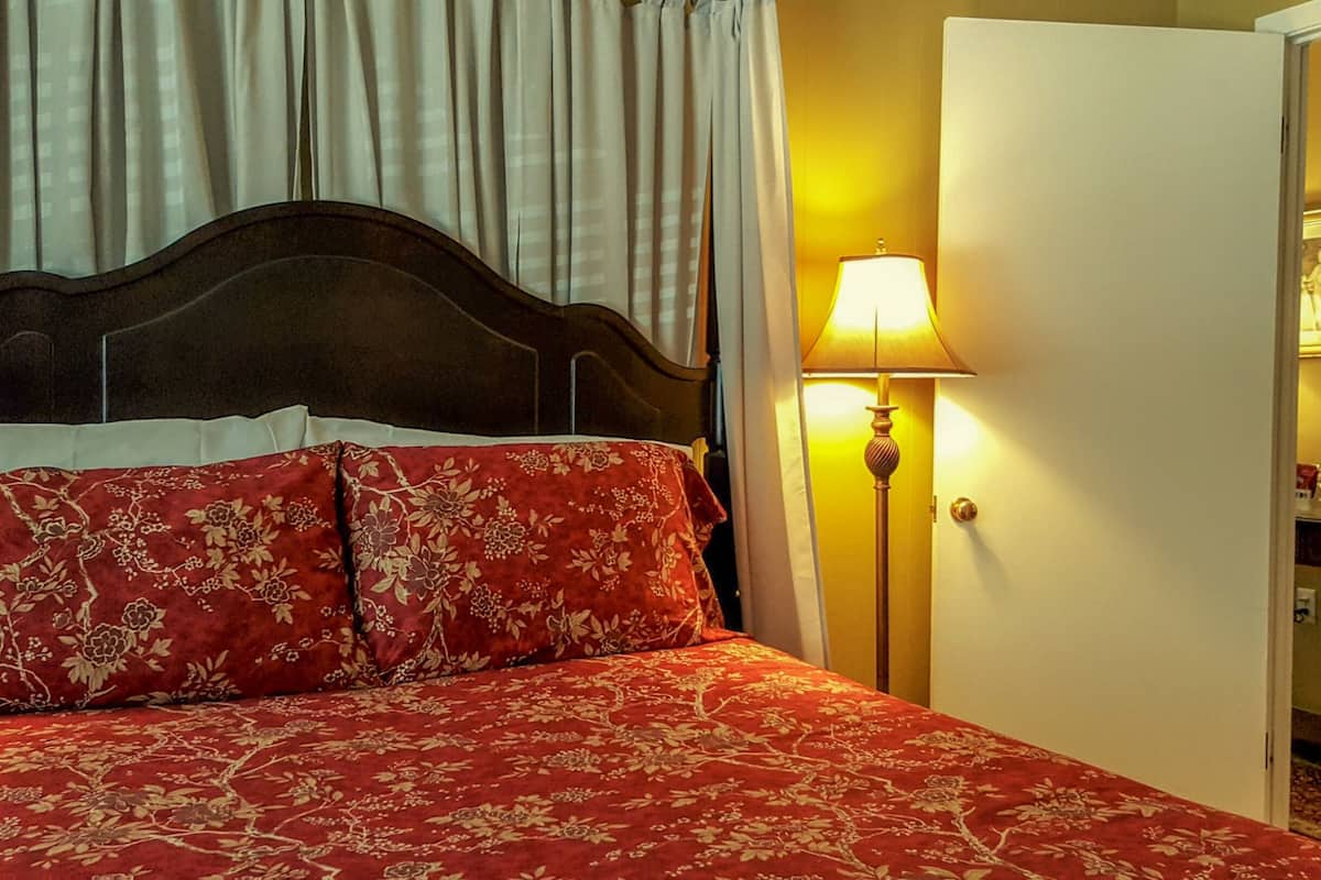 A hotel room with a bed, curtains, a lamp, a painting, and a small table with glasses.