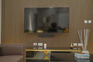 Ein modernes Wohnzimmer mit einem flachbildfernseher, der an einer Holzwand befestigt ist, einem Glastisch mit geometrischem Design und einem braunen Sofa.