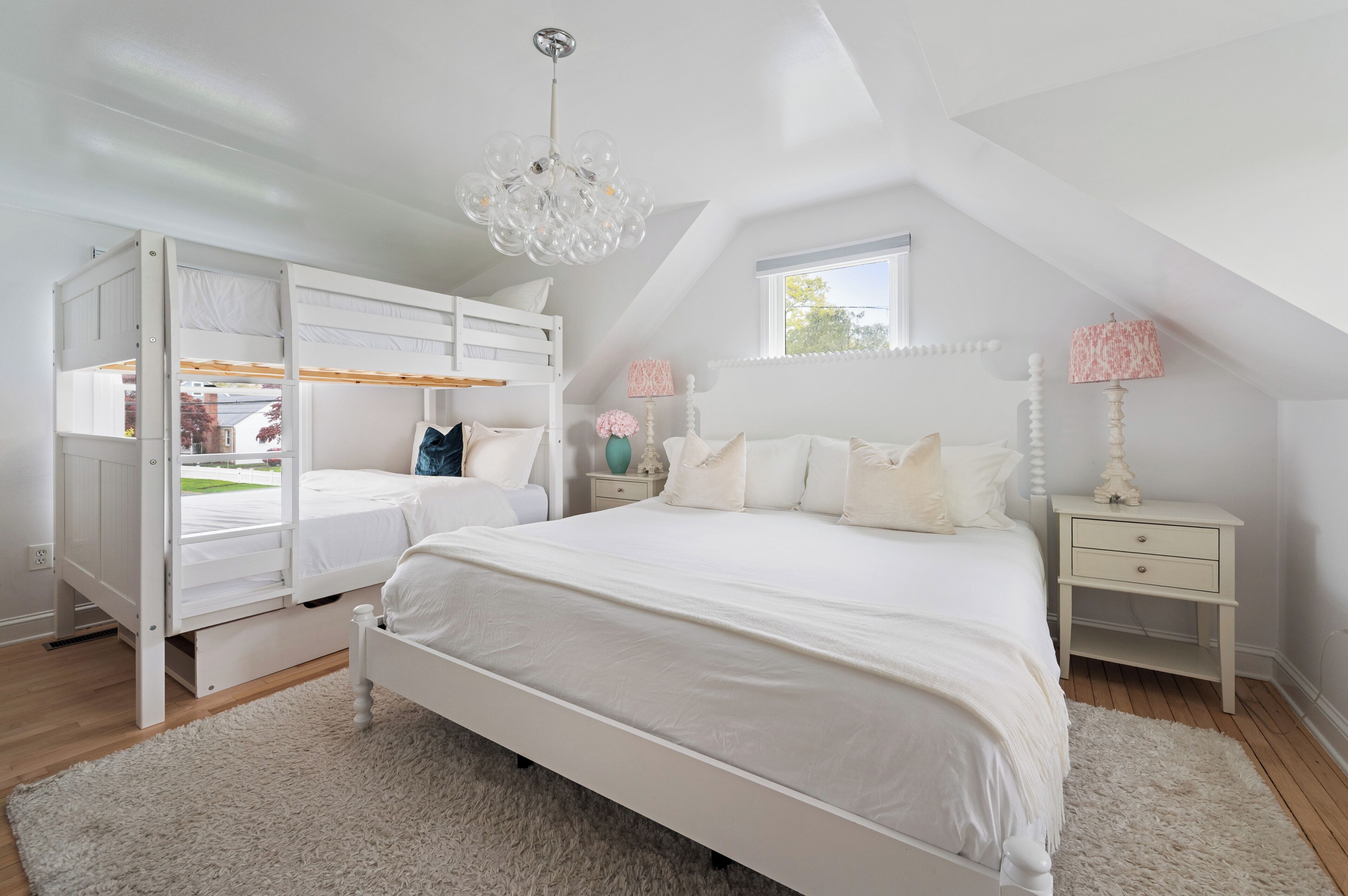Un dormitorio con una cama litera, una cama grande, un candelabro, una ventana y una mesita de noche con una lámpara.