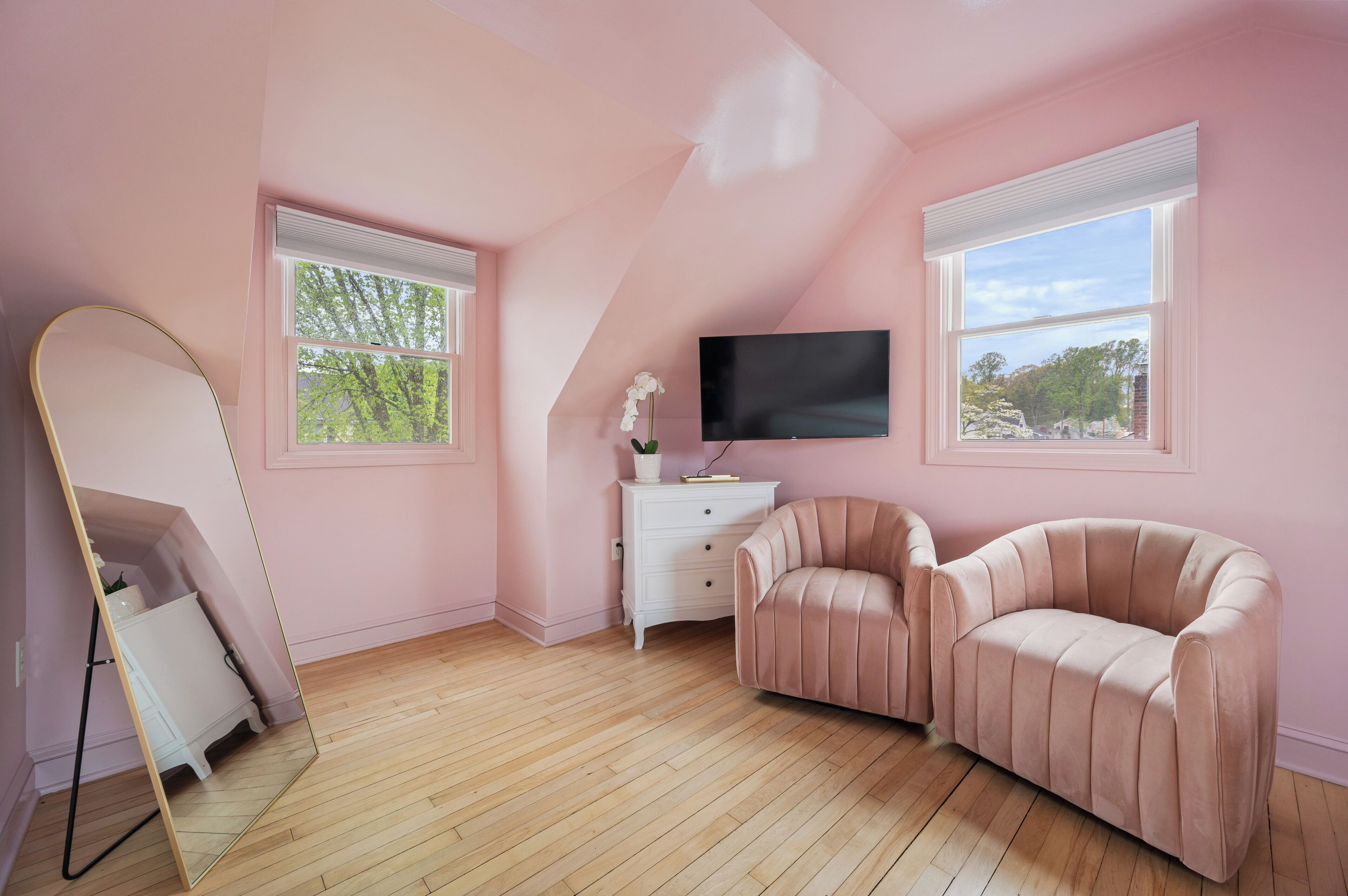Une pièce décorée dans des tons roses, avec un téléviseur à écran plat, une commode blanche, un canapé rose et un grand miroir.