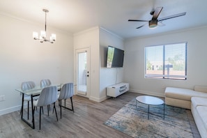 A modern living room with a dining area, a ceiling fan, a flat-screen TV, and a large window with blinds. - San Portella 2119 (Tempe)
