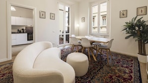 Un salon moderne avec un canapé, une table à manger et des chaises. On aperçoit une cuisine au fond, et une grande fenêtre offre une vue sur les bâtiments.