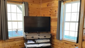 Um quarto de cabana acolhedor, com paredes de madeira, uma janela grande, uma TV de tela plana, um sofá e um tapete estampado.