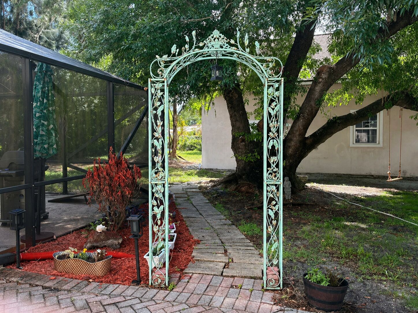 Een tuinpad met een sierlijk metalen bogenpoort, een bankje en bloempotten.