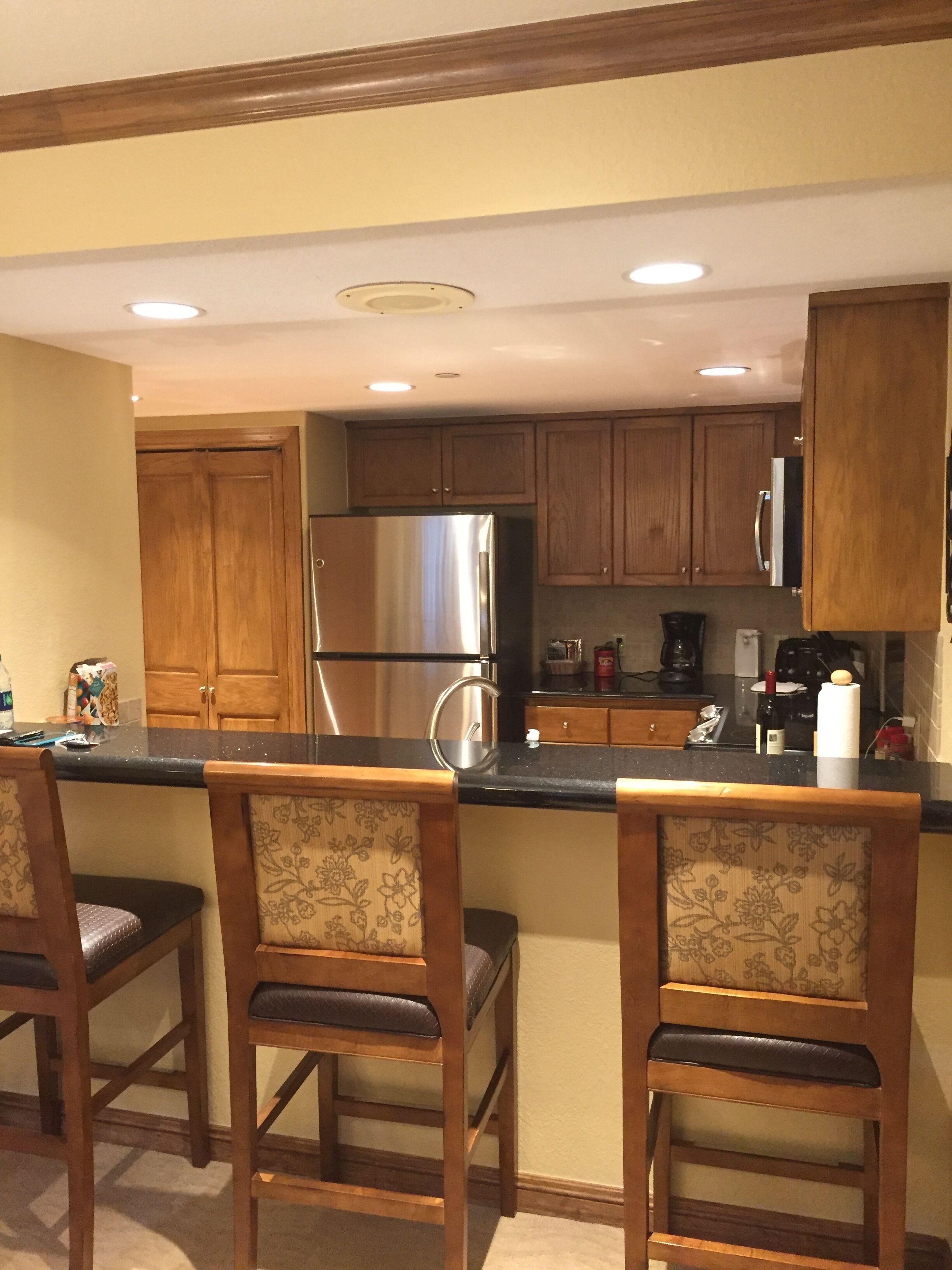 A kitchen with wooden cabinets, a stainless steel refrigerator, a coffee maker, and two upholstered bar stools.