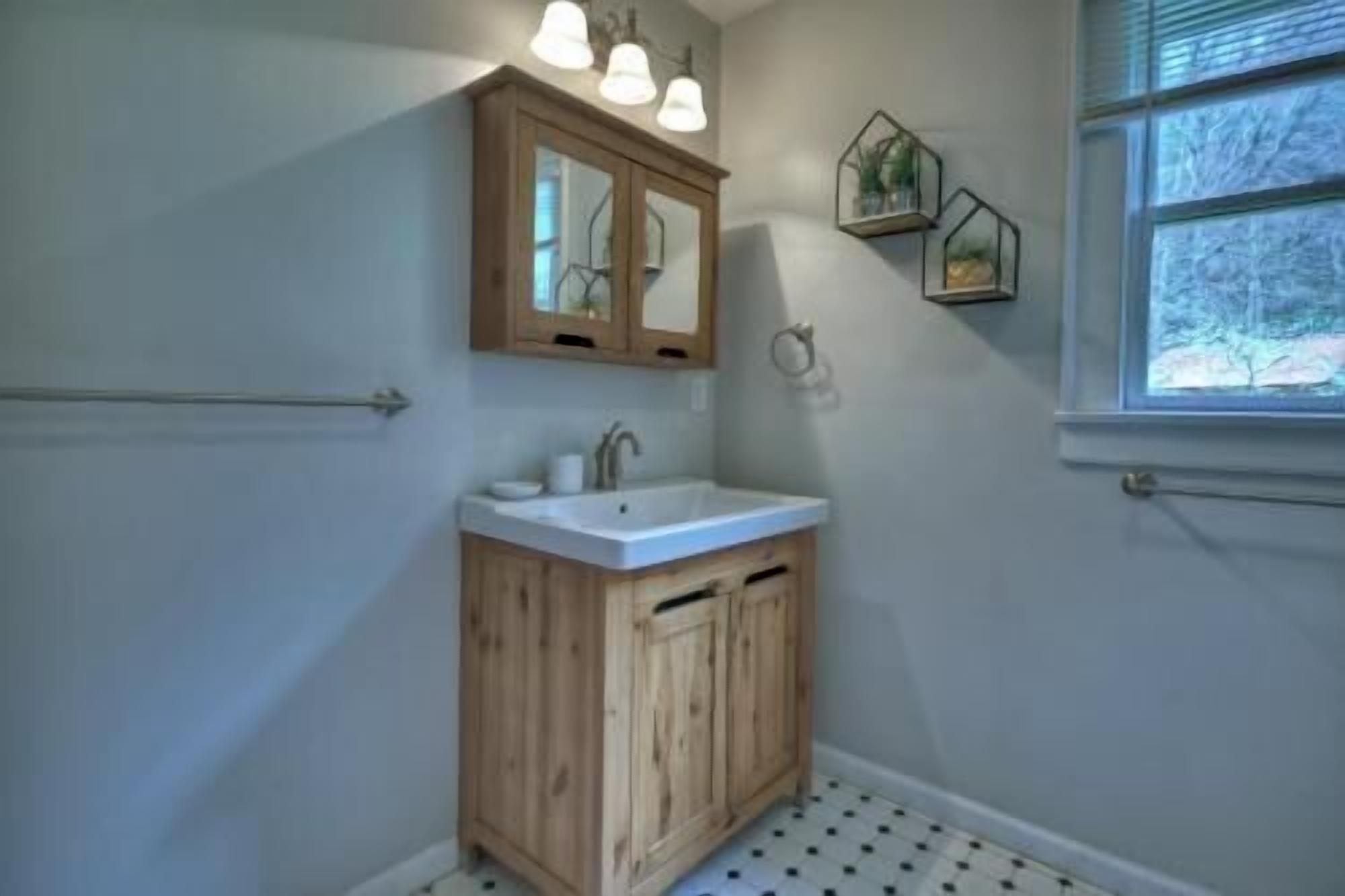 A bathroom with a wooden vanity, a white sink, and a mirror above it. There are two wall-mounted lights above the mirror and a window with blinds to the right.